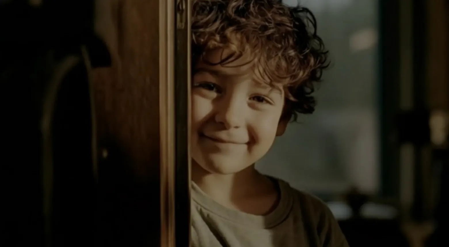 A young child with curly brown hair and a big smile peeks out from behind a door, softly lit by natural light coming from the side.